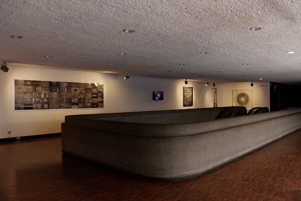 A modern interior gallery or lounge space with low, curved upholstered seating in the foreground, wooden flooring, and framed abstract artworks mounted on the walls under recessed ceiling lights.