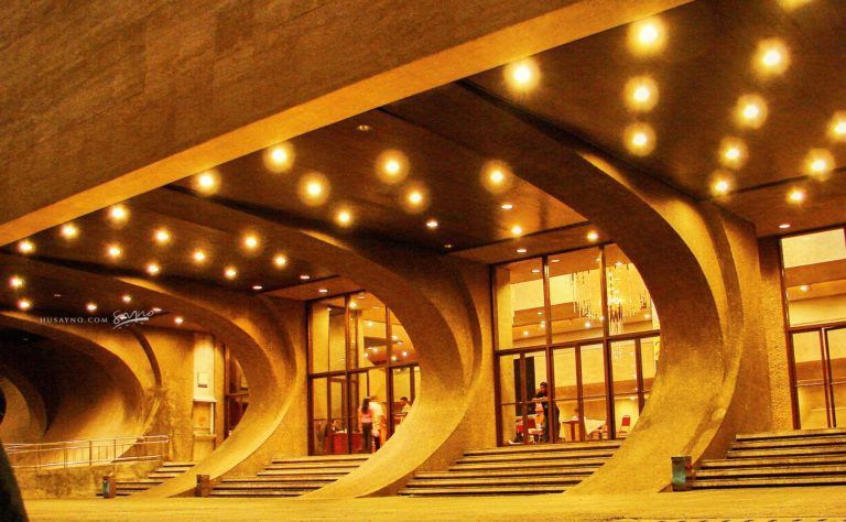 A dramatic architectural building entrance with large curved concrete arches, warm amber lighting, wide steps, glass doors, and a few small people visible near the entrance.