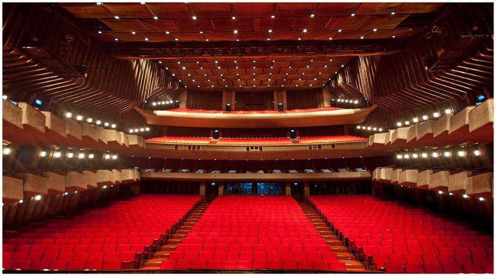An empty large auditorium or theater viewed from the stage area, with rows of bright red seats, tiered balconies, wood-paneled walls, and many ceiling lights illuminating the hall.
