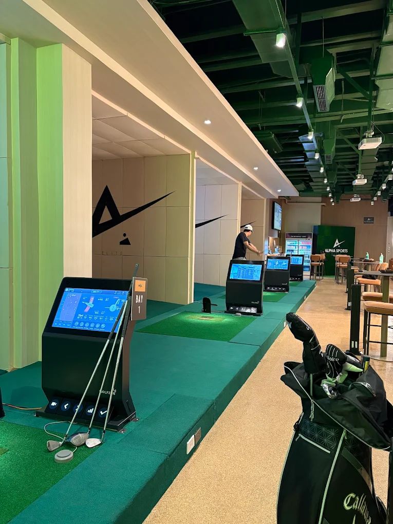 Row of indoor golf simulator bays in a modern sports lounge, each with a hitting mat, screen, and control monitor; a golfer is practicing in one bay, golf clubs and a bag sit near the front, and signage reads "BAY 1" and "ALPHA SPORTS."