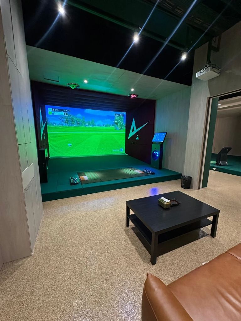 Private indoor golf simulator room with a large projected golf course screen, central hitting mat, wall-mounted lighting, and a small touchscreen kiosk to the right; a low black coffee table sits in the lounge area outside the bay, with a leather sofa partially visible in the foreground.