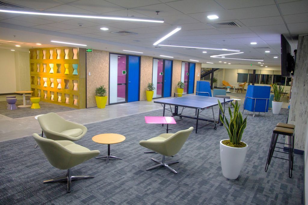 Modern open-plan office lounge with carpeted seating area, colorful small tables and chairs, a ping-pong table, potted plants, and glass meeting-room doors; bright ceiling lights and geometric wall decor create a casual coworking space.