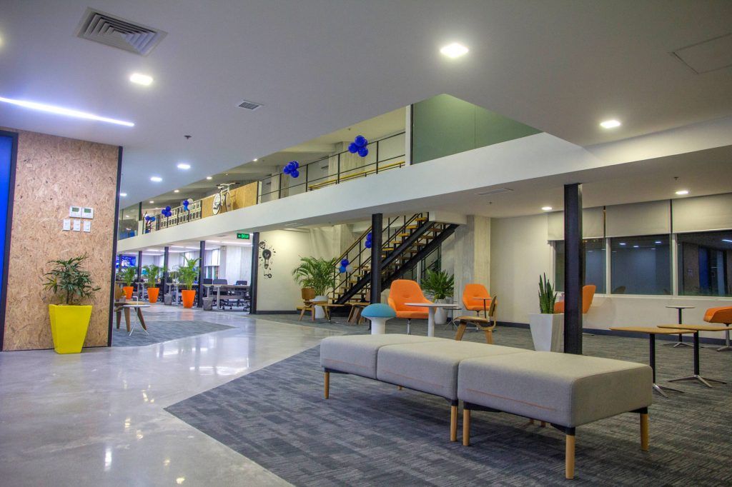Spacious contemporary office common area with a central staircase, long corridor, colorful lounge chairs and tables, potted plants, and a large low bench; bright overhead lighting and glass-walled rooms suggest a coworking or corporate workspace.