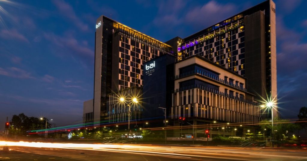 Night exterior of a contemporary hotel building with illuminated windows and the text "bai HOTEL" visible on the facade, with light trails from passing traffic in the foreground.