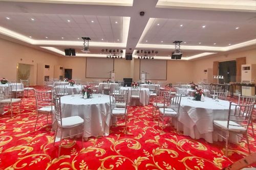 An indoor banquet hall set up for an event, with round tables covered in white tablecloths, silver chairs, and a bright red patterned carpet. The room has a stage area with large projection screens, ceiling lights, and a formal reception layout; no readable text is visible.