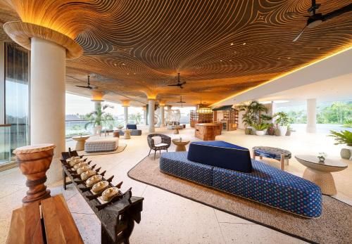 Luxury resort lounge interior with a sculpted wooden ceiling, white columns, woven chairs, sofas, decorative drums, indoor plants, and a bright open-air tropical setting.