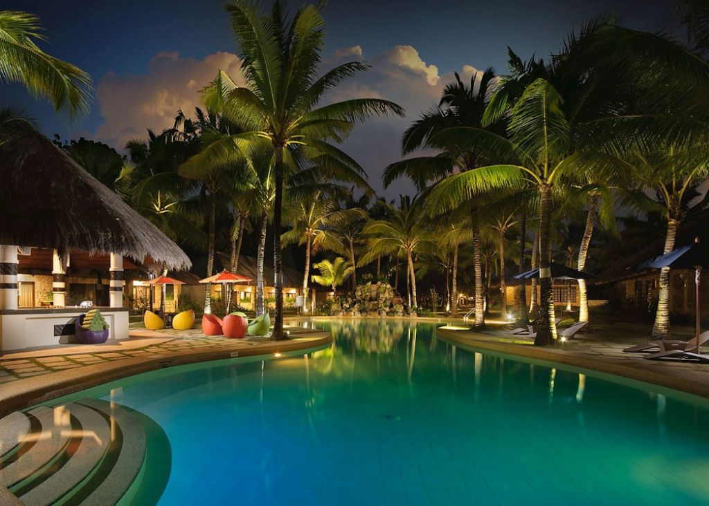 Nighttime tropical resort pool area with glowing turquoise water, palm trees, thatched-roof structures, colorful seating, and ambient landscape lighting.