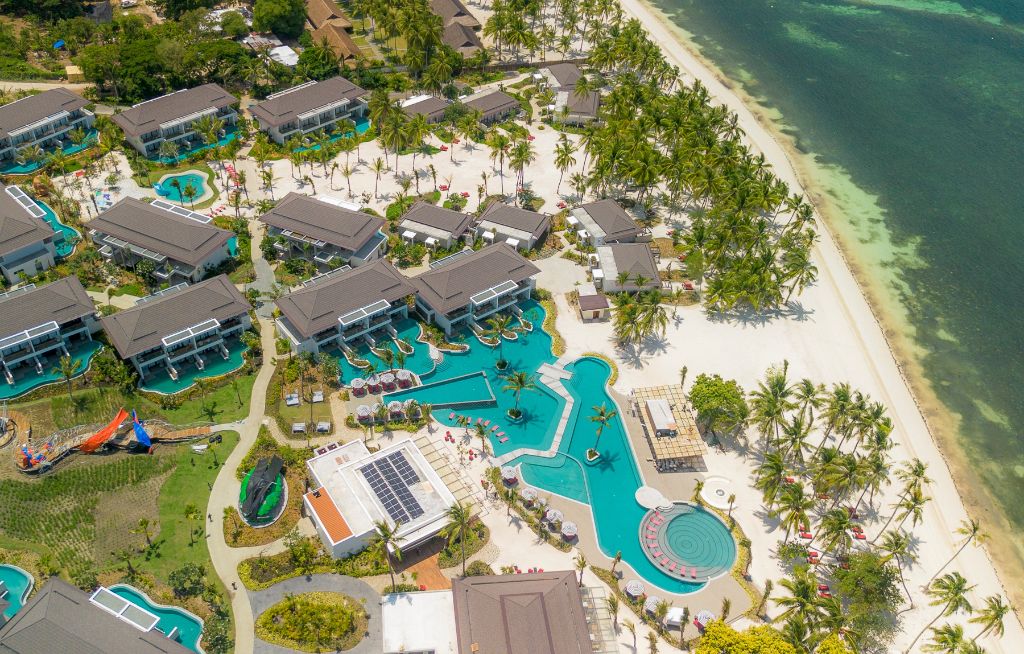 Aerial view of a beachfront resort complex with multiple villas, winding pools, palm-lined white sand beach, and clear ocean water along the shore.